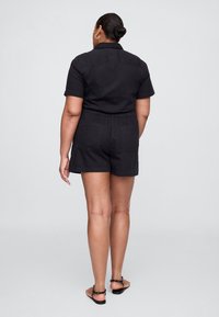 Mono corto de manga corta negro hecho de tela con cuello, cintura elástica y dos bolsillos traseros. Se lleva con sandalias planas sencillas.