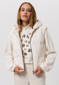 Lichtgewicht crèmekleurig jack met capuchon, ritssluiting en elastische manchetten. Draagt over een wit shirt met gouden luipaardvlekken.