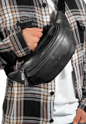 Marsupio in pelle nera con un design curvo, dotato di chiusura con zip e piccoli dettagli in metallo, indossato sopra una camicia a quadri.