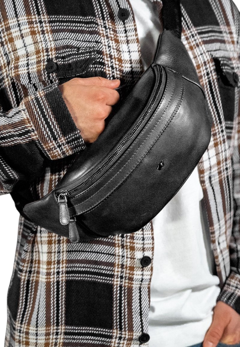 Marsupio in pelle nera con un design curvo, dotato di chiusura con zip e piccoli dettagli in metallo, indossato sopra una camicia a quadri.