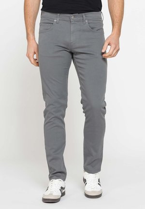 Uomo vestito con pantaloni grigi aderenti e sneakers bianche in piedi davanti a uno sfondo chiaro e uniforme.
