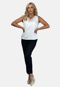 Biała bezrękawnikowa bluzka z teksturowym dekoltem, w zestawieniu z dopasowanymi czarnymi spodniami i złotymi espadrylami. Gładkie tło.