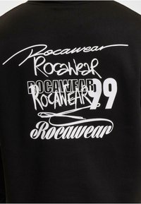 Crna dukserica s bijelim otisnutim tekstom. Dizajn uključuje više varijanti riječi "Rocawear" u različitim fontovima, veličinama i stilovima.