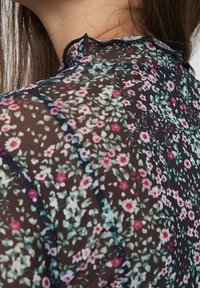 Blusa floreale trasparente con sfondo nero, caratterizzata da piccoli motivi floreali rosa e verdi, delicato scollo a giorno e tessuto leggero.