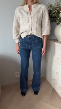 Žena u krem pletenom kardiganu s dugmadima, plavim širim trapericama i crnim cipelama, stoji unutra pored cvjetnog vaza na radnoj površini.