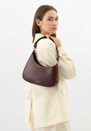 Vrouw in een crèmekleurig pak met een donkerbruine leren schoudertas met gouden hardware en het "wolas" logo.