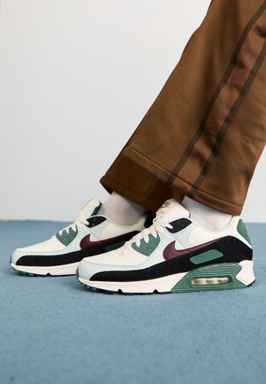 Les baskets Nike Air Max 90 présentent un dessus en cuir et en mesh blanc avec des accents vert foncé, bordeaux et noir. Semelle en caoutchouc texturé, unité Air visible.