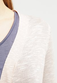 Světle růžový pletený cardigan s výstřihem do V, zdobený texturovaným vzorem, nošený přes modré tričko.