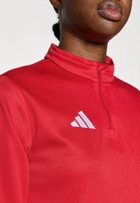 Κόκκινο αθλητικό τοπ με φερμουάρ μέχρι τη μέση, κατασκευασμένο από λείο ύφασμα. Διαθέτει ψηλό γιακά και λευκό λογότυπο στο αριστερό στήθος. Εφαρμοστό σχέδιο.