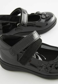 Zapatos de charol negro con punta redondeada, banda de Velcro y un lazo decorativo en la parte frontal. Plantilla texturizada con un patrón floral.