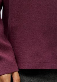 Burgundské pletené svetr vyrobený z měkké tkaniny, se žebrovanými manžetami a spodním lemem, jemně texturovaný povrch, kombinovaný se šedou tkaninou pod ním.