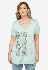 Camiseta verde menta con escote en V, detalles de encaje decorativos y dobladillo curvado. Presenta un estampado gráfico con lentejuelas plateadas y un detalle de lazo.