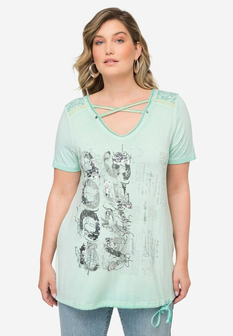 Camiseta verde menta con escote en V, detalles de encaje decorativos y dobladillo curvado. Presenta un estampado gráfico con lentejuelas plateadas y un detalle de lazo.