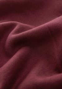 Bordeauxrode stof met een textuurweefsel, die zacht valt. Heeft subtiele schaduwen en highlights, die diepte en beweging benadrukken.