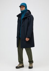 Giacca impermeabile della marina con cappuccio sopra un fleece teal, abbinata a pantaloni oliva e scarpe robuste. Presenta una vestibilità comoda e un design funzionale.