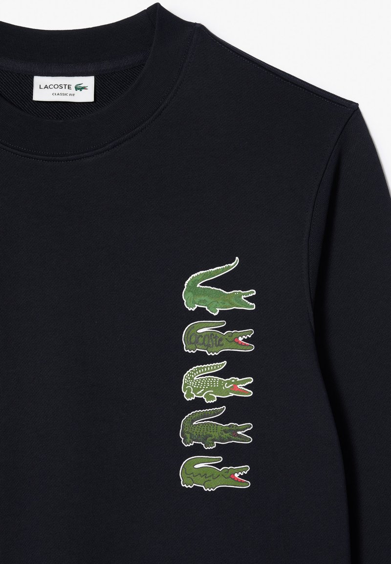 Sudadera negra con una línea vertical de cinco logotipos de cocodrilo verdes con acentos rojos. Corte clásico con un cuello redondo.