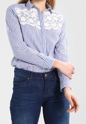 Femme portant une chemise à rayures verticales bleues et blanches avec un détail en dentelle blanche sur la poitrine et un jean bleu foncé.