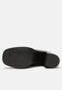 Suela de zapato negra con una superficie texturada, que presenta franjas horizontales y una ligera curvatura. El material parece gomoso con un acabado mate.