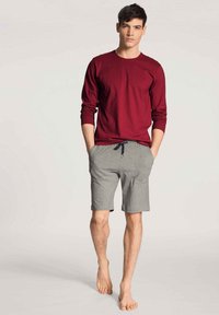 Rode longsleeve shirt gecombineerd met lichtgrijze shorts met koordjes. Zacht materiaal, losse pasvorm en blote voeten. Eenvoudig ontwerp zonder patronen.