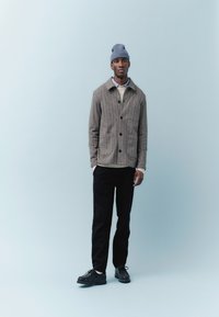 Uomo con berretto grigio, giacca marrone con bottoni sopra maglione crema, pantaloni neri e scarpe nere, in piedi davanti a uno sfondo azzurro pallido.