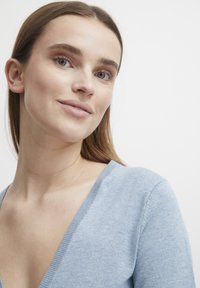 Pull en tricot bleu clair avec un décolleté en V. Texture douce, style ajusté, avec des accents côtelés subtils au niveau du col et des manches.