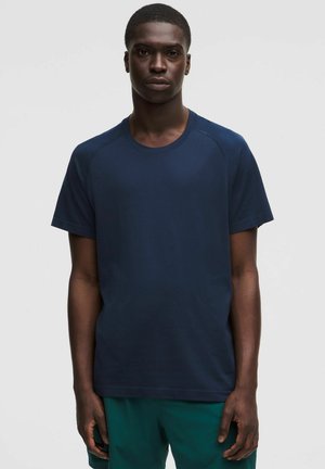 Uomo con capelli corti che indossa una maglietta blu navy a maniche corte e pantaloni verdi, in piedi davanti a uno sfondo chiaro e uniforme.