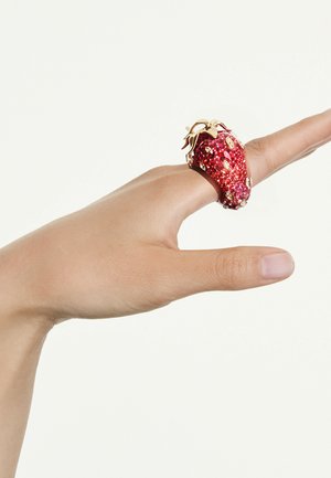 Hand mit großem, rotem, glitzerndem ring in Form einer Erdbeere am Mittelfinger vor einfachem weißem Hintergrund.