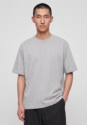 Homme aux cheveux courts noirs portant un t-shirt uni gris clair à manches courtes et un pantalon noir, debout devant un fond clair uni.