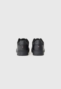 Paire de baskets basses noires vues de dos avec un petit logo rectangulaire sur chaque talon sur un fond clair uni.