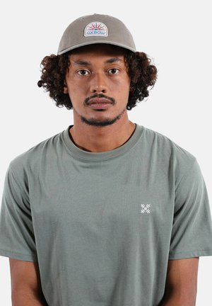 Homme aux cheveux bouclés portant une casquette grise avec un logo de soleil et un t-shirt vert olive avec un petit emblème blanc sur la poitrine.