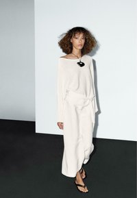 Let og off-white sweater med bred halsudskæring og lange ærmer, parret med løse off-white bukser og sorte sandaler. Sort vedhængs halskæde.