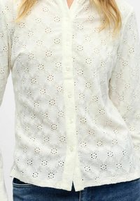 Camicia bianca con bottoni e pizzo floreale indossata con jeans blu, che mostra una fine trama del tessuto e parziali capelli biondi.
