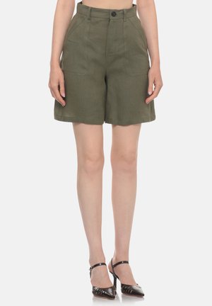 Donna con pantaloncini verde oliva a vita alta con tasche frontali e tacchi neri con cinturino alla caviglia e punta appuntita, in piedi davanti a uno sfondo monocromatico.