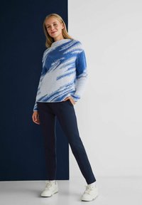 Maglione morbido blu e bianco con un motivo ondulato, abbinato a pantaloni blu navy e sneakers bianche. Il modello è posizionato davanti a uno sfondo scuro.