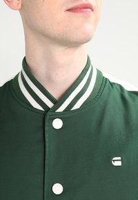 Närbild av en person som bär en mörkgrön jacka med vitrandig ribbad krage och vita tryckknappar.