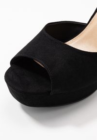 Lahjan lähikuva tyylikkäästä mustasta korkeasta korkokenkästä, jossa on suede-pinta ja avonaiset varpaat, neutraalia taustaa vasten.