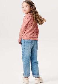 Dziewczyna w różowej, wzorzystej koszuli z długim rękawem oraz jasnoniebieskich, podwiniętych dżinsach, w połączeniu z beżowymi butami. Włosy falowane i luźno opadające.