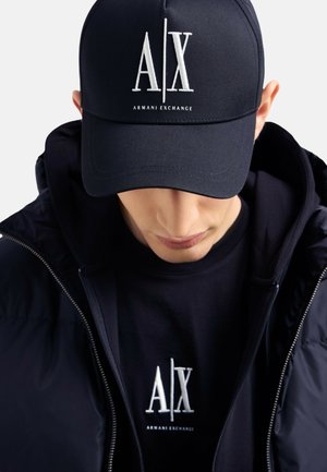 Cappellino da baseball blu navy con logo bianco "A/X", indossato con un felpa nera e una giacca, che mette in mostra un design elegante e dettagli in tessuto texture.