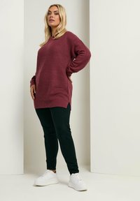 Geflochtenes bordeauxrotes Sweatshirt mit langen Ärmeln und seitlichen Schlitzen, kombiniert mit schwarzen Slim-Fit-Hosen und weißen Sneakers, vor einem neutralen Hintergrund.