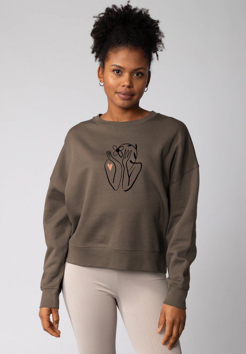 Olijfgroene, cropped sweatshirt van zacht materiaal met een zwarte lijntekening van handen en een bloem, vergezeld van een roze hartaccent.