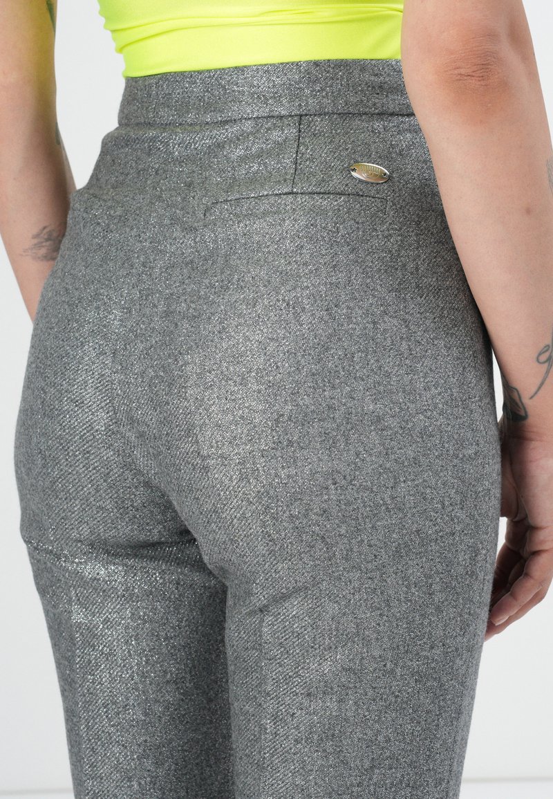 Persona vestida con pantalones plateados brillantes y una camiseta amarilla neón, con tatuajes visibles en los brazos, vista trasera enfocando en la cintura y las caderas.