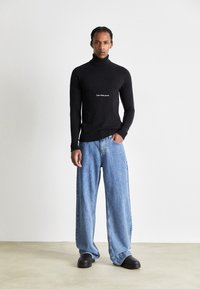 Maglione nero a collo alto con polsini a coste, con la scritta bianca "Calvin Klein Jeans" sul petto. Indossato con jeans a gamba larga blu chiaro e scarpe nere.