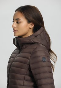 Braune Pufferjacke mit horizontalen Steppnähte, hohem Kragen und einem Reißverschluss. Mit einem runden Logo-Akzent am linken Ärmel.