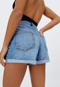 Primo piano di una persona che indossa pantaloni shorts in denim azzurro chiaro arrotolati e una maglietta nera, vista di spalle con una mano sulla vita.