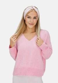 Maglione rosa a scollo a V in un materiale morbido e soffice, con maniche lunghe. Indossa una fascia rosa coordinata, con i capelli lisci parzialmente sciolti.