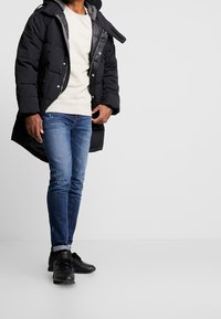 Veste matelassée noire avec doublure grise, capuche et boutons-pression ; sweat-shirt crème en dessous ; jean bleu avec détails subtils ; chaussures de sport noires.