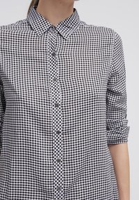 Camicia a scacchi bianca e nera con colletto, apertura frontale con bottoni, maniche arrotolate e una texture del tessuto liscia.