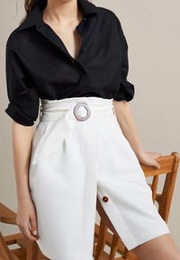 Donna che indossa una camicia nera a maniche lunghe infilata in pantaloni corti bianchi a vita alta con una cintura di tessuto e una fibbia circolare.