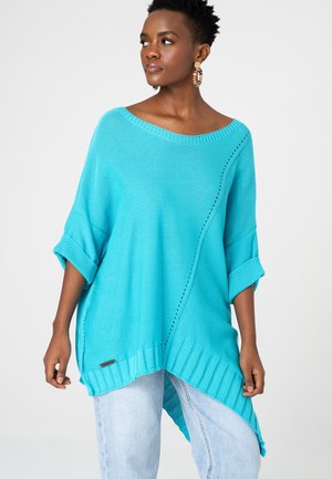 Kobieta ubrana w luźny turkusowy dziergany sweter z podwiniętymi rękawami i jasnoniebieskie jeansy, patrząca w lewo.