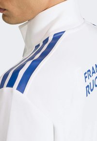 Biała sportowa kurtka z niebieskimi pionowymi paskami na ramionach i napisem „FRANCE RUGBY” w kolorze niebieskim na plecach. Gładka faktura.